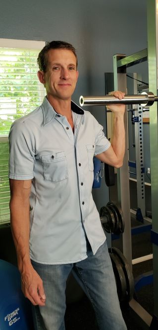 Picture of Scott Cline the owner of Unique Physique, smiling holding onto the 45lb barbell bar, on the gym floor.